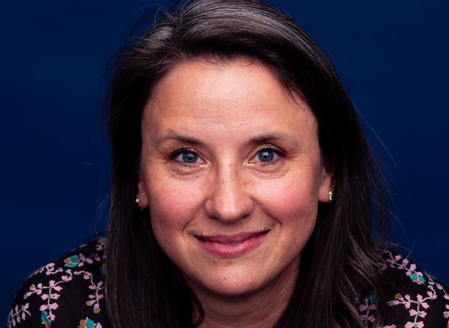 Photograph of a white woman smiling into the camera, portrait-style. She's got long brown hair and wears a flotal patterned sleeveless. 